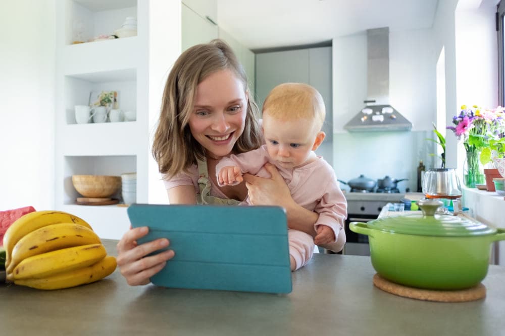 A parent holding an infant while looking at a tablet in a kitchen